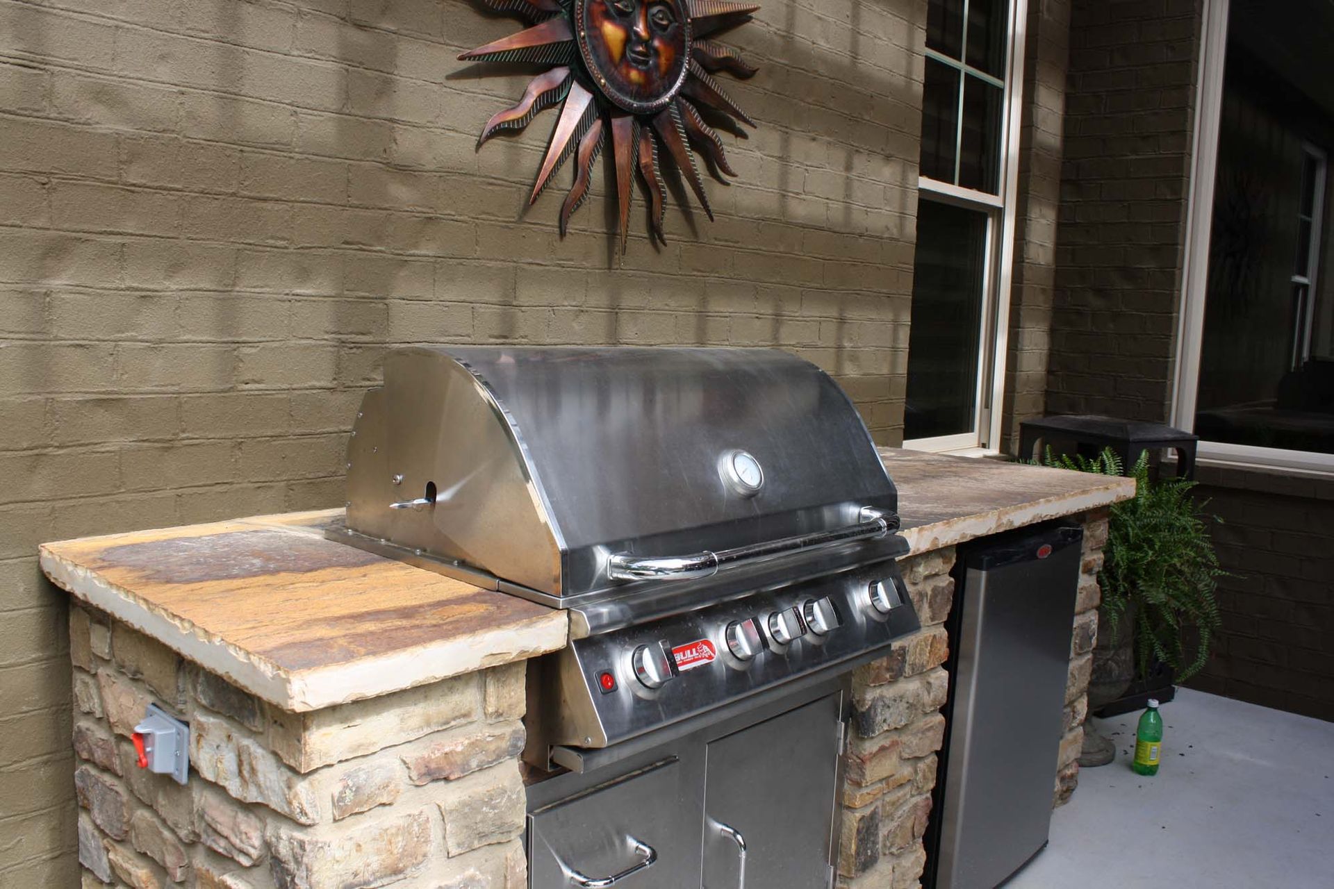 Outdoor stainless steel gas grill beside a stone countertop and trash bin against a brick wall.