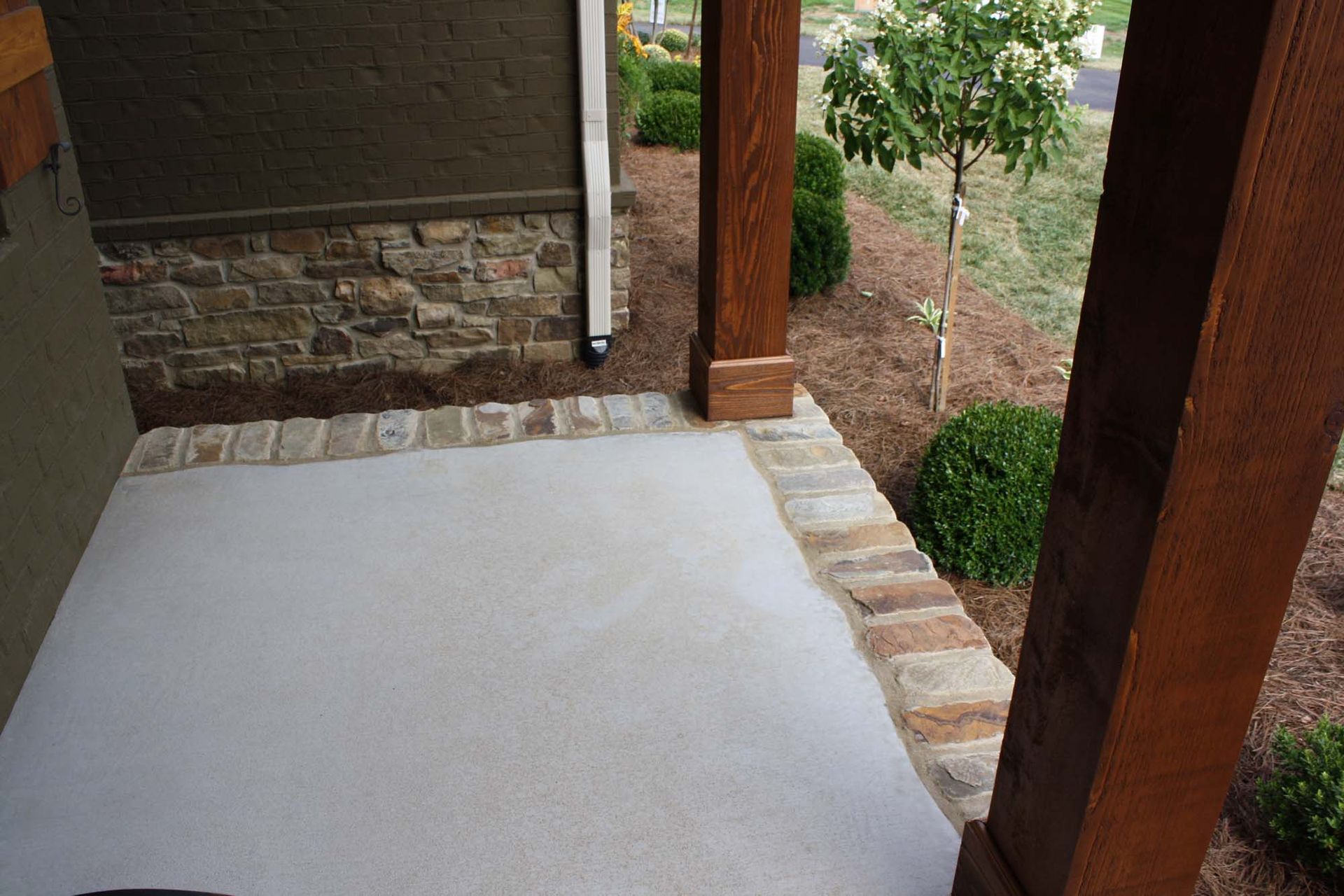Concrete porch floor beside brick edging and wooden posts, with a landscaped yard beyond.