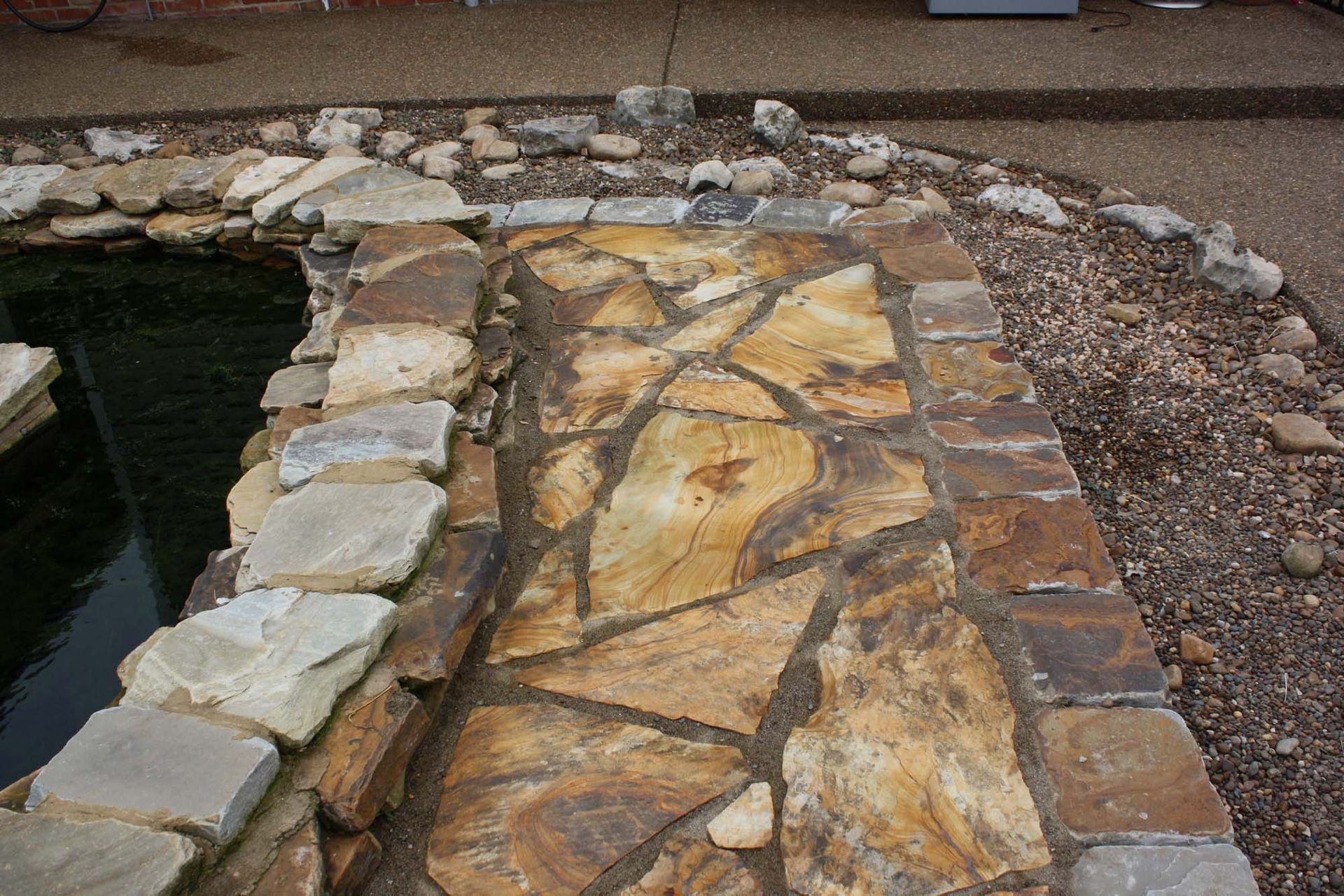 Stone walkway beside a pond, edged with rocks and bordered by gravel