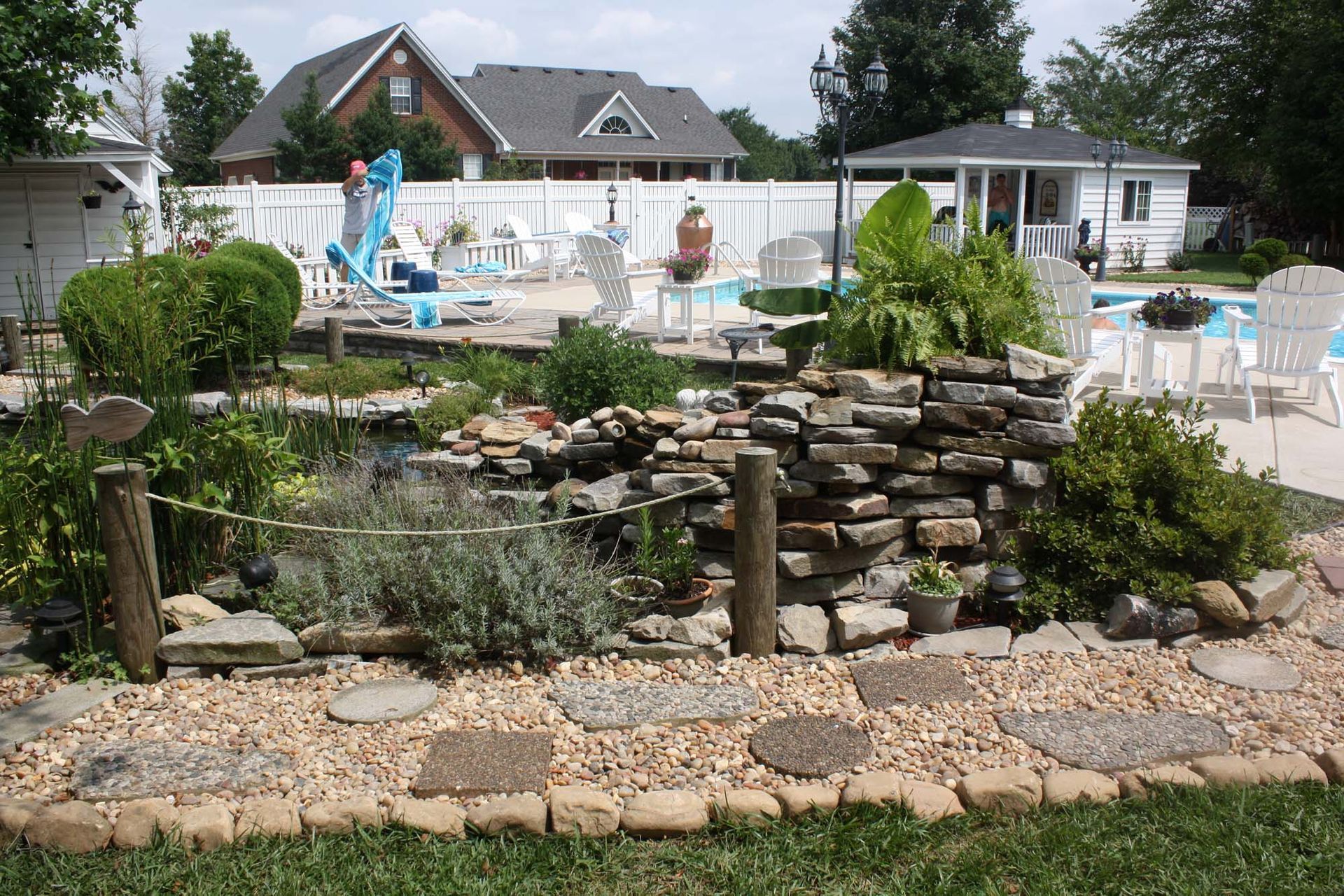 Backyard pool with stone garden, patio chairs, white fence, and a house in the background