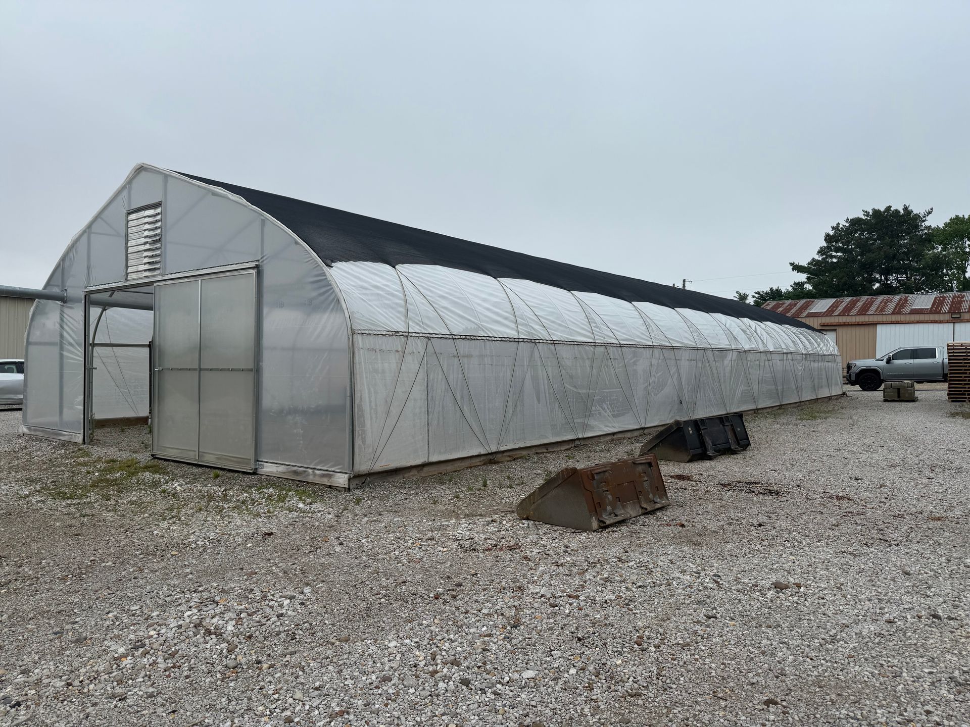 Small greenhouse tunnel with translucent plastic panels on a gravel lot under an overcast sky