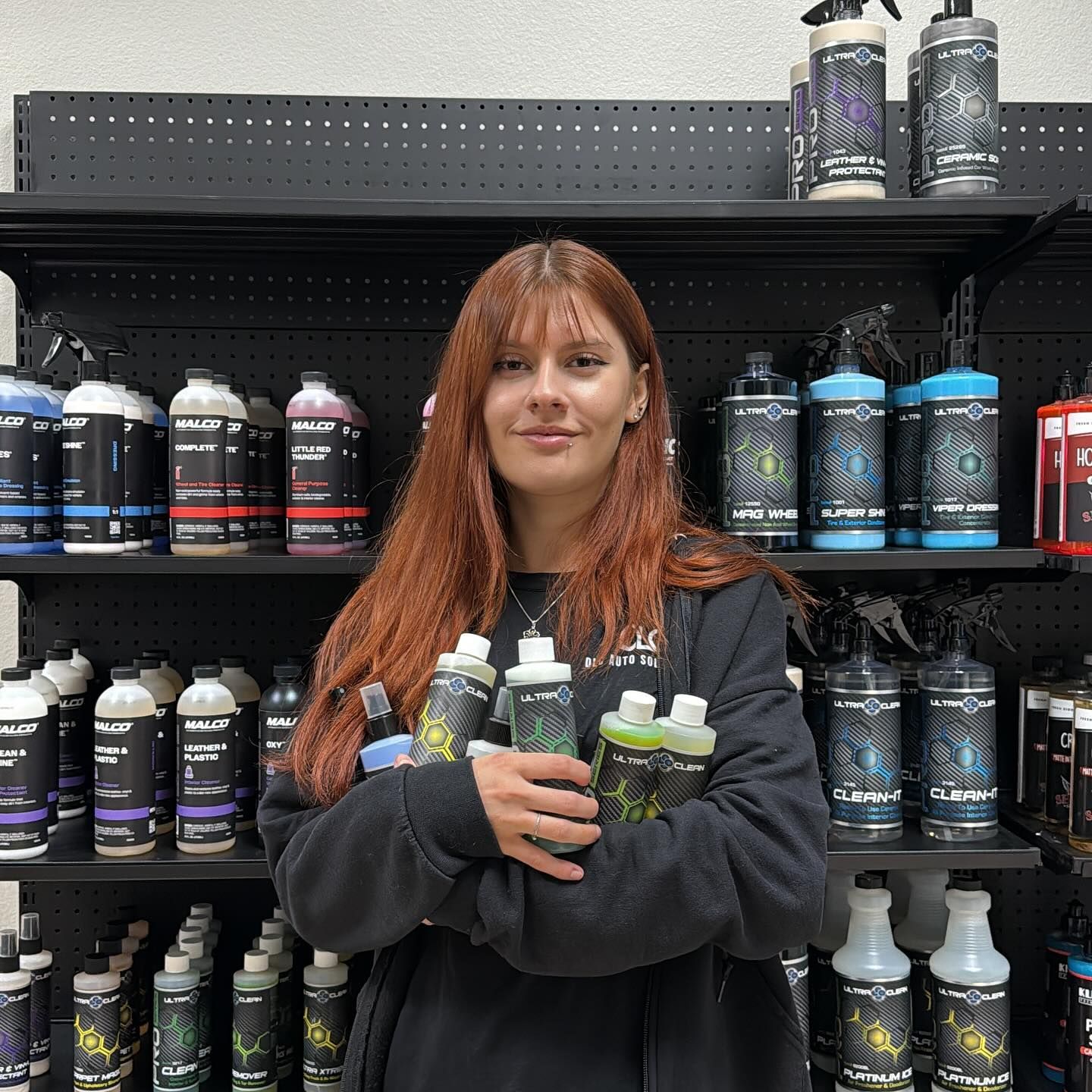 Woman holding cleaning product bottles in a shop, arms crossed. Products on shelves behind her.