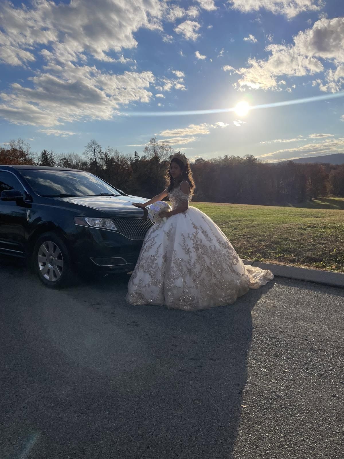 Woman in a white ball gown standing beside a black SUV on a road at sunset.