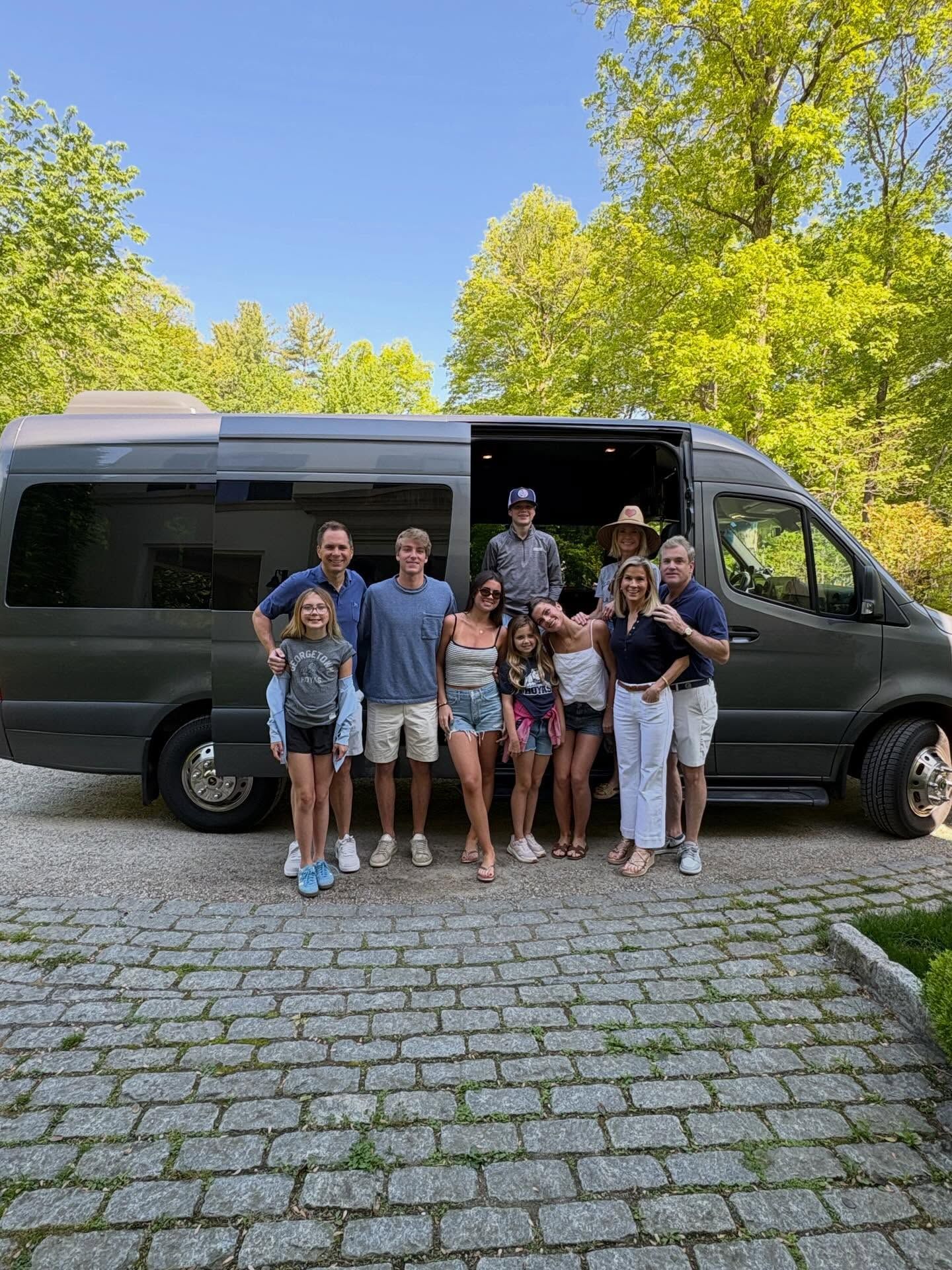 Family group posing beside a dark van on a cobblestone road with bright green trees in the background