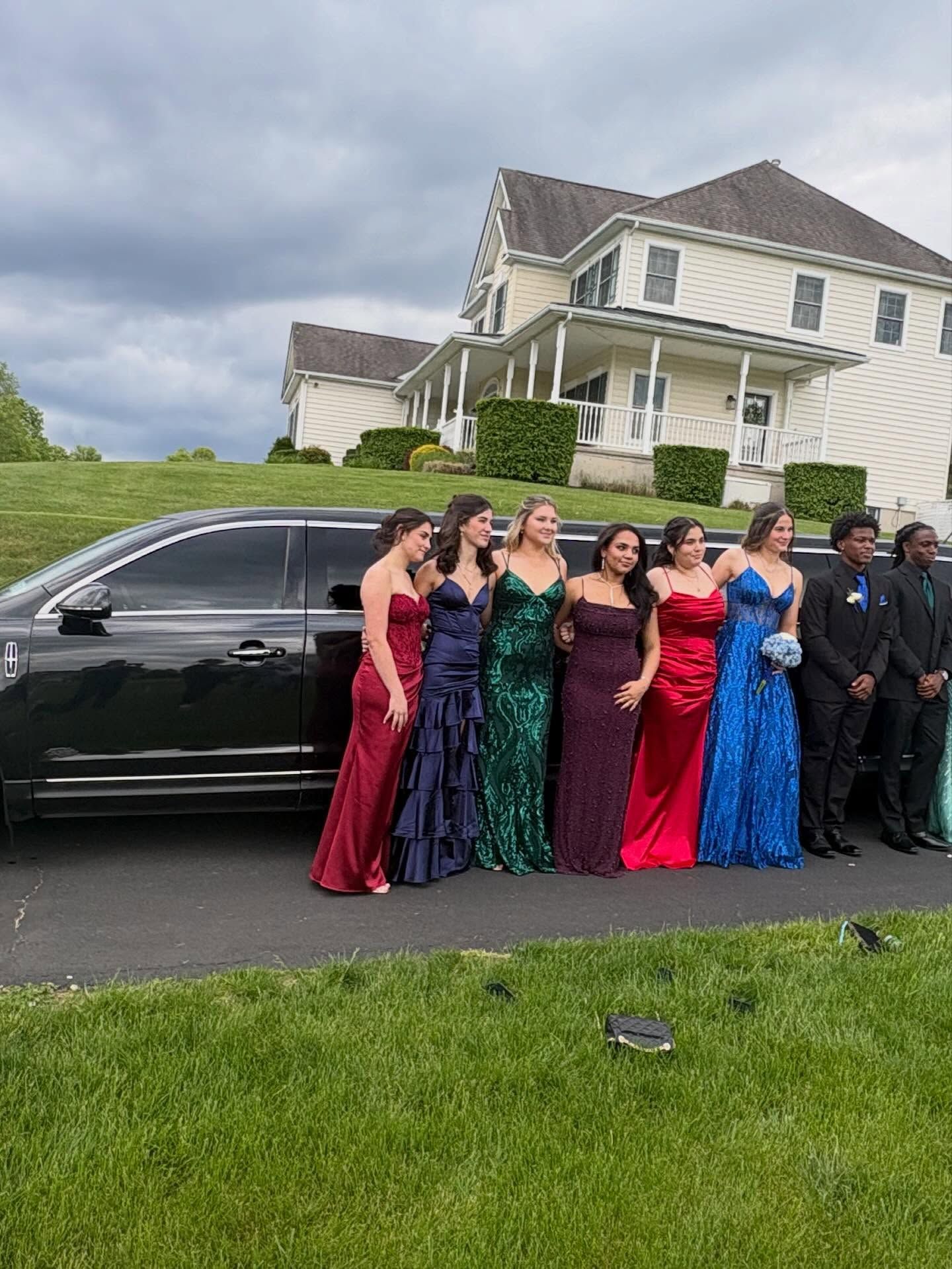 Group in formal dresses poses beside a black limousine in front of a large house.