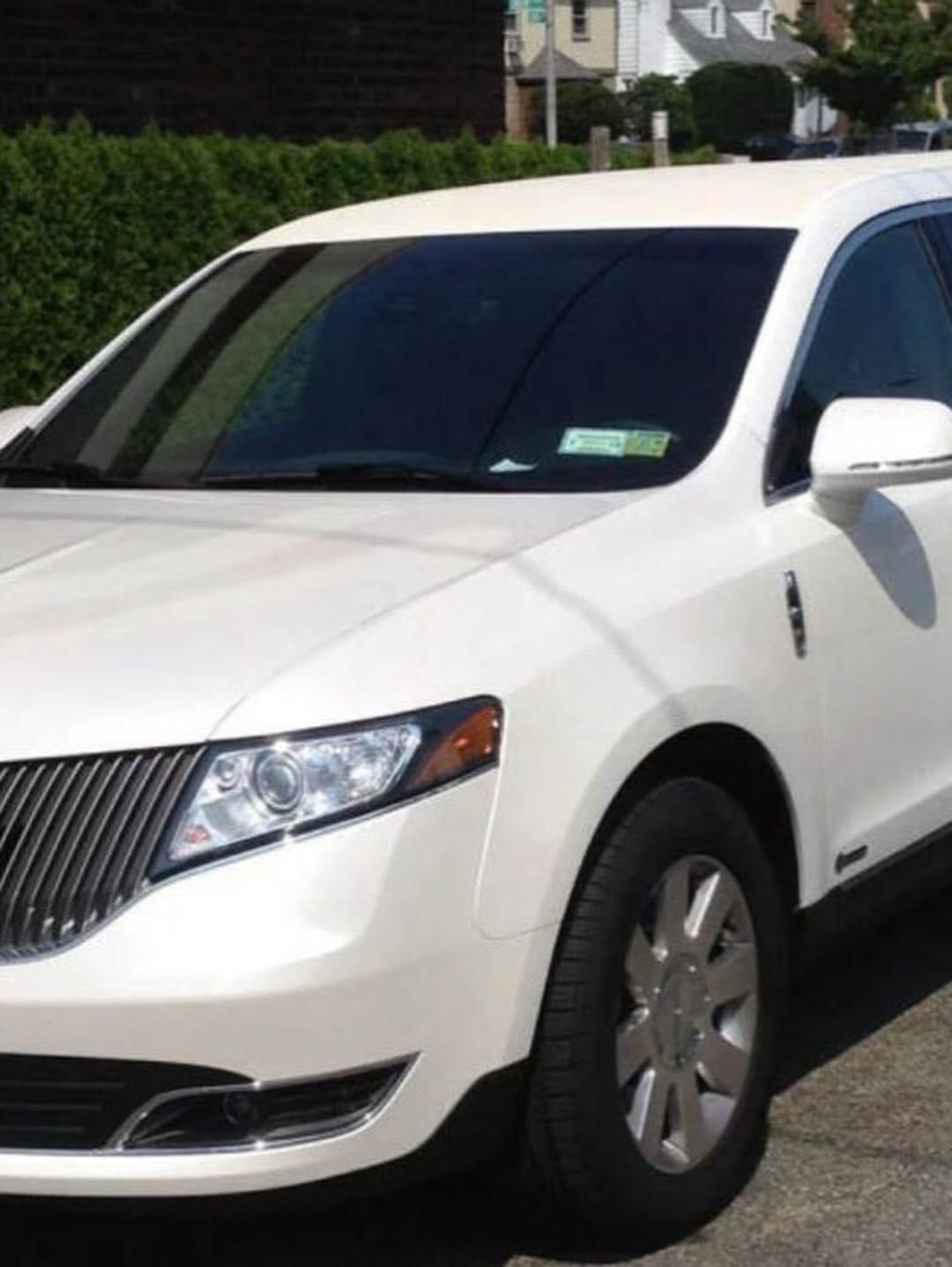 White sedan parked outdoors in front of bushes and a house, front-left view
