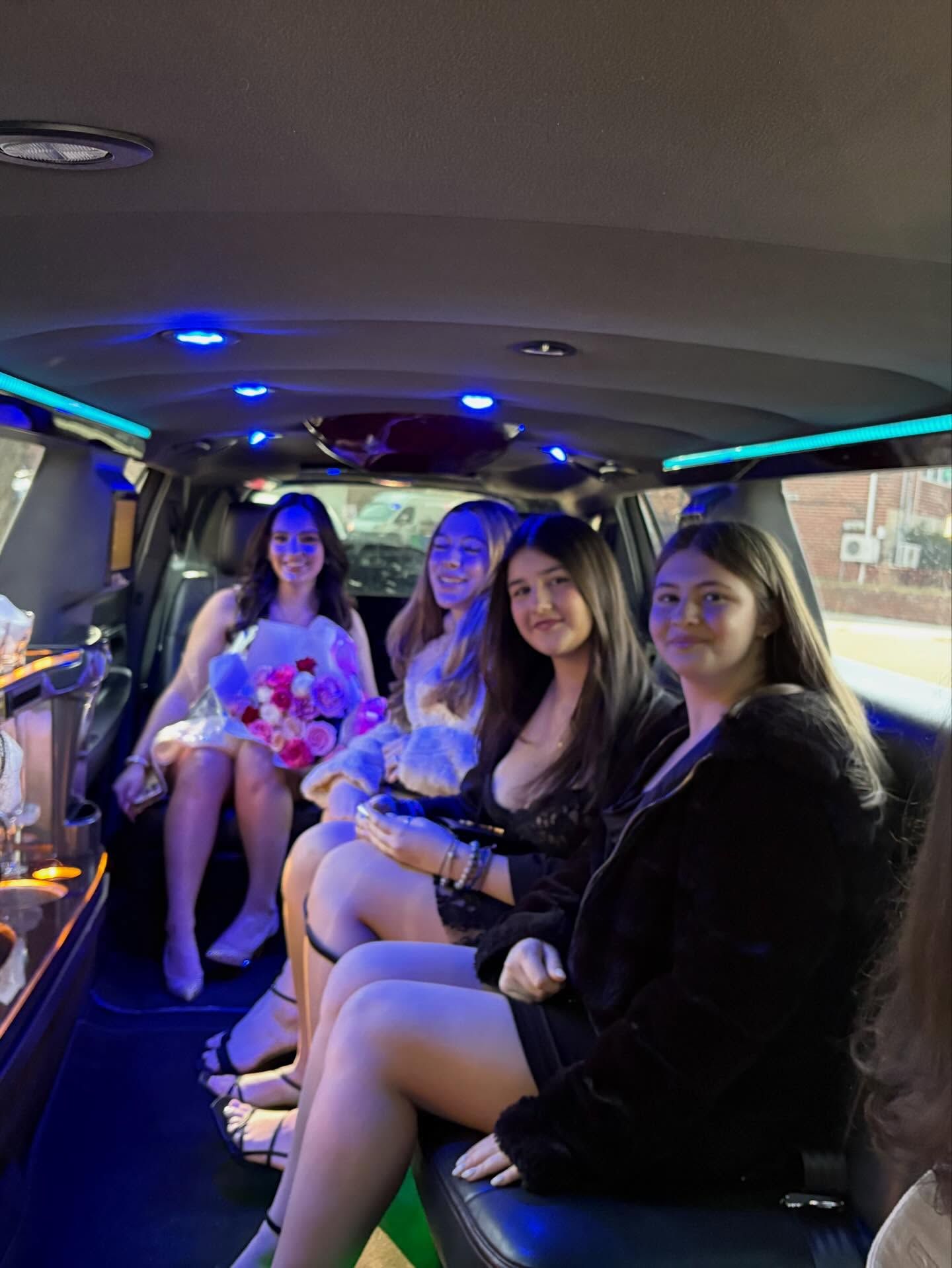 Four women seated in a limousine, lit with blue accent lights, smiling inside the vehicle.