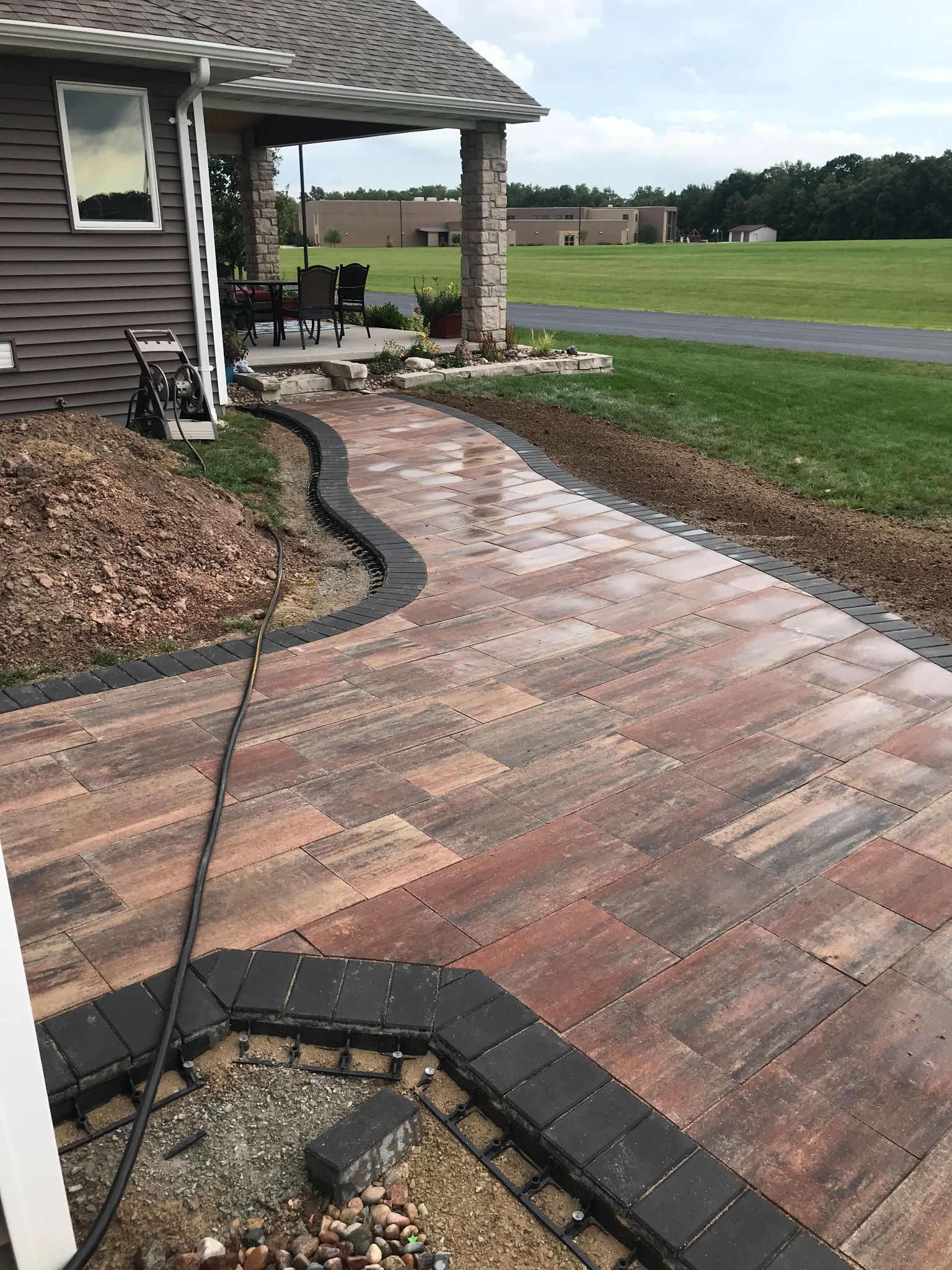 Brick walkway with a black border leading beside a house to a grassy yard and open field