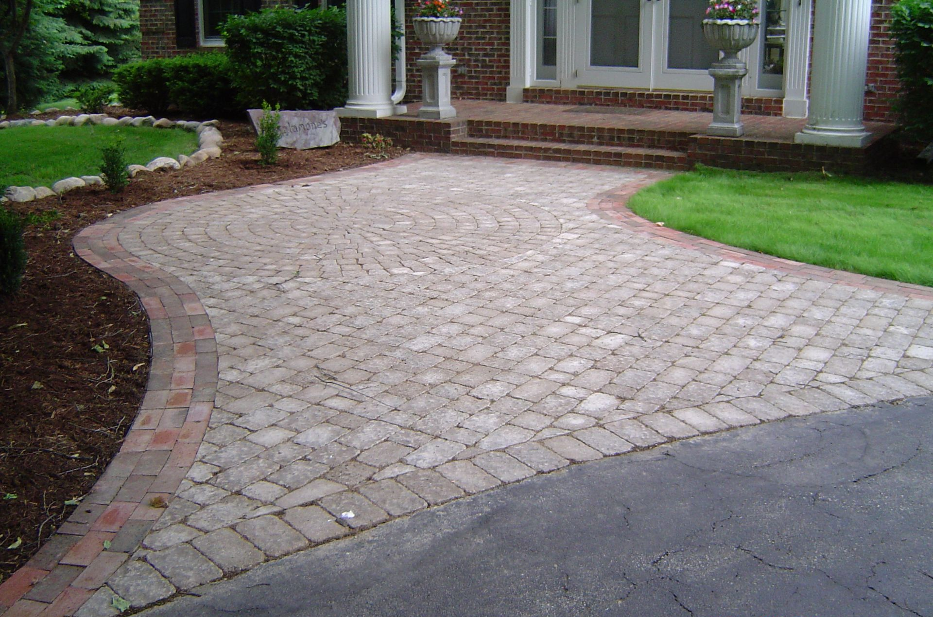 Brick-paved driveway curves to a house entrance with white columns and green landscaping.