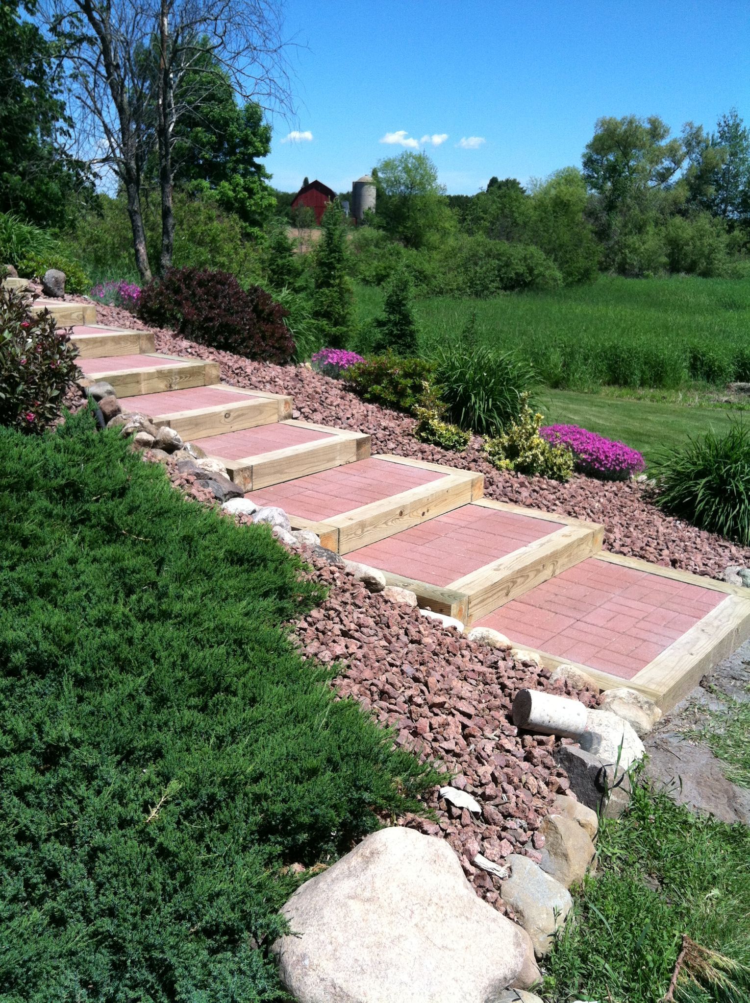 Stone steps in a landscaped garden with green shrubs, pink flowers, and a blue sky.