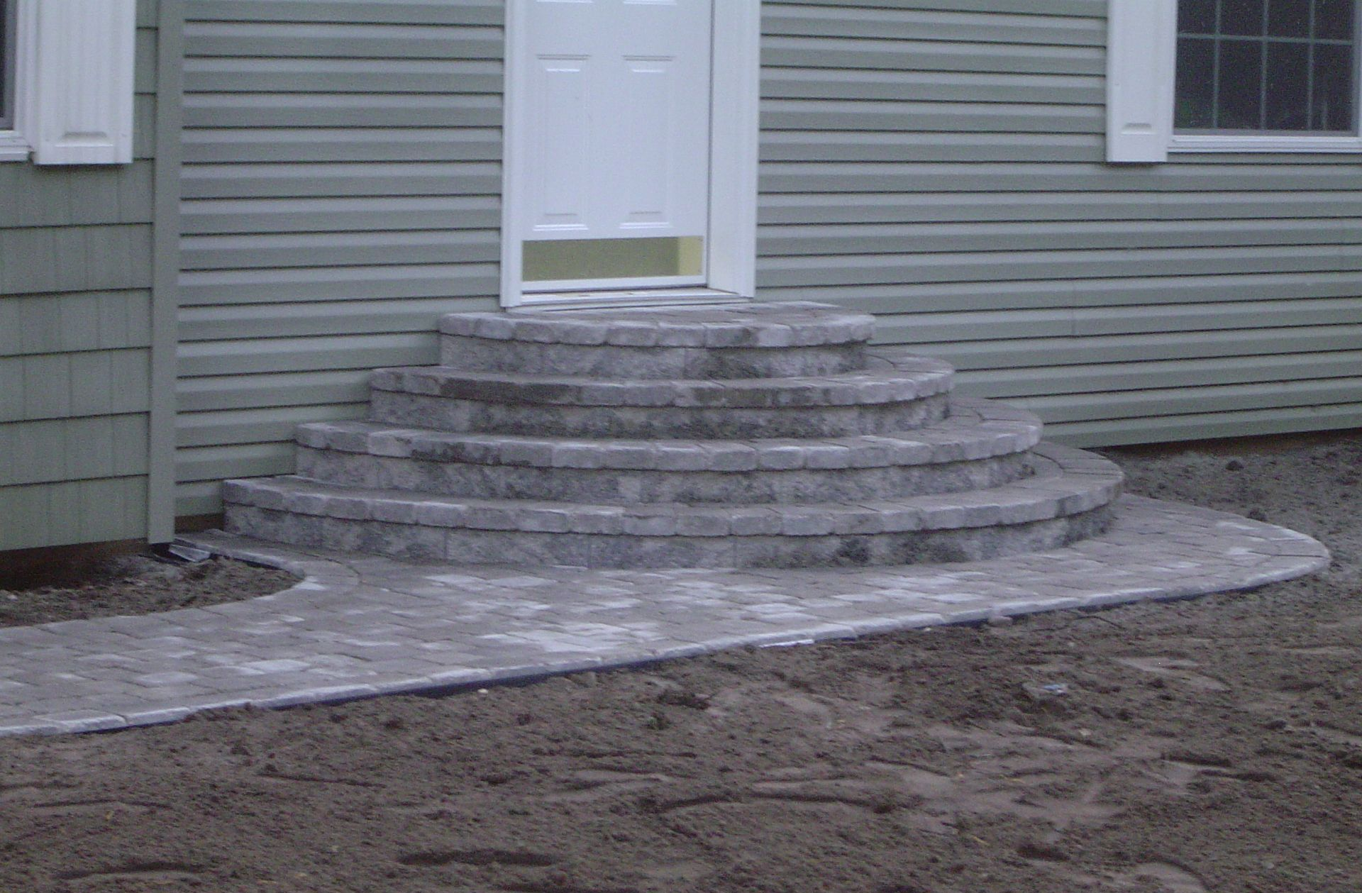 Concrete front steps leading to a white door on a gray-sided house, with a bare dirt yard.