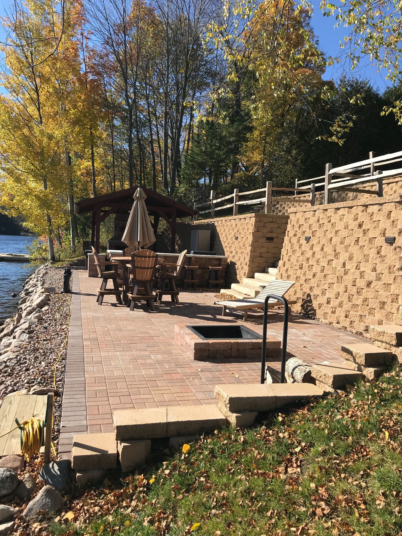 Brick patio with outdoor chairs beside a stone wall, riverbank, and autumn trees in sunlight