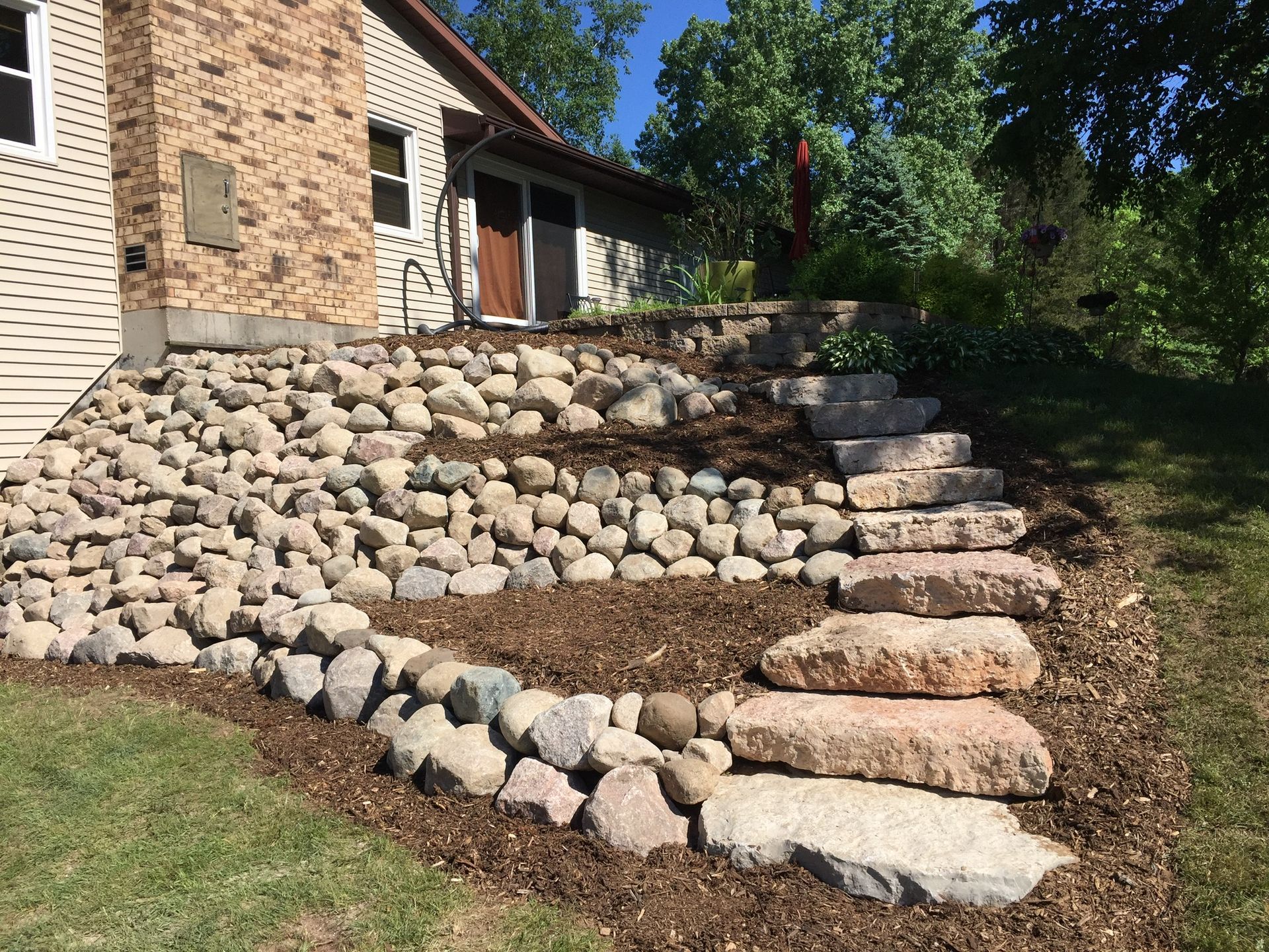 Stone retaining wall with steps beside a house, landscaped with rounded river rocks and grass.
