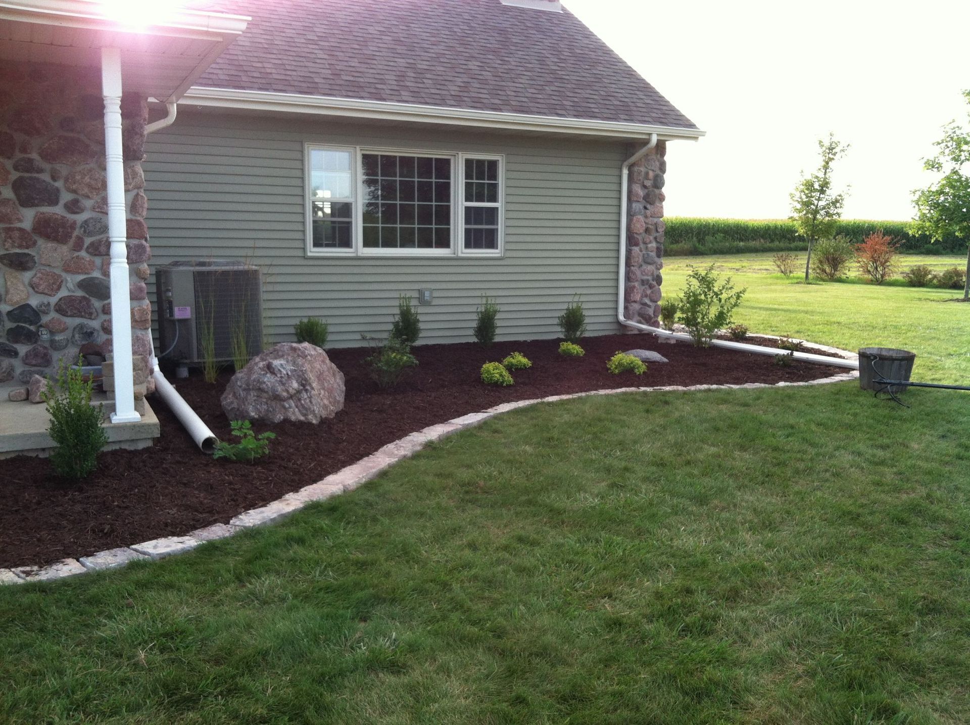 Landscaped backyard with green lawn, mulched flower bed, shrubs, and side of a house with stone and siding