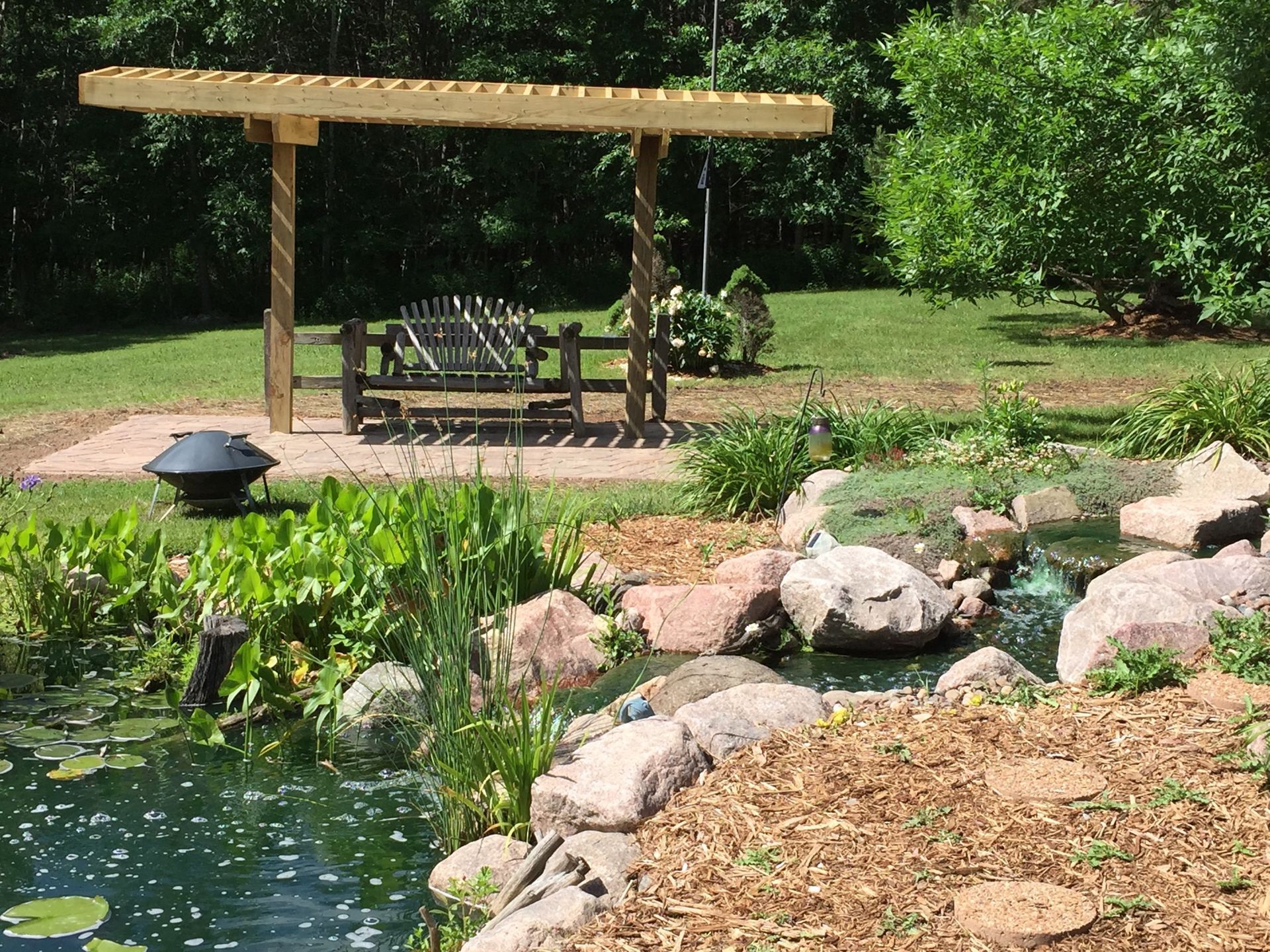 Backyard garden with pond, rocks, wooden bench under a pergola, and lush greenery