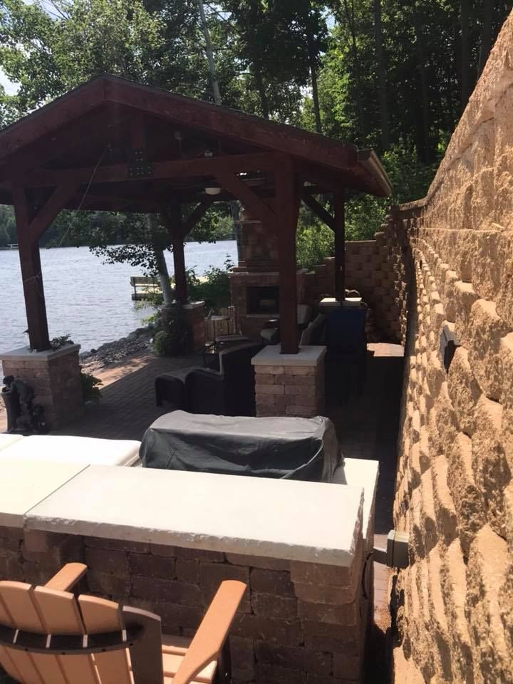 Lakeside patio with a wooden gazebo, tables, and seating beside a stone wall and water.