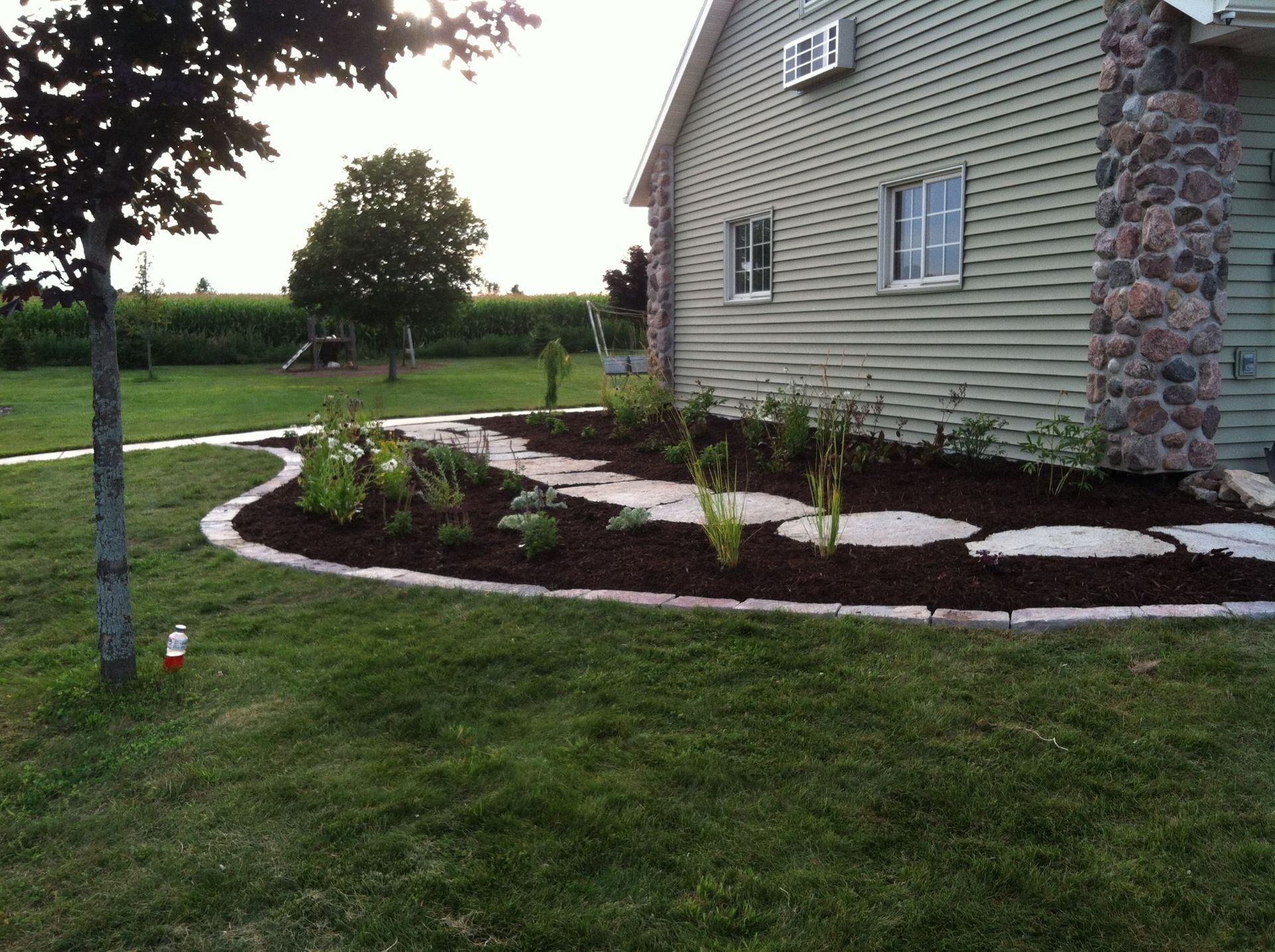 Curved landscaped flower bed beside a stone-and-siding house with fresh mulch and a small tree in the yard