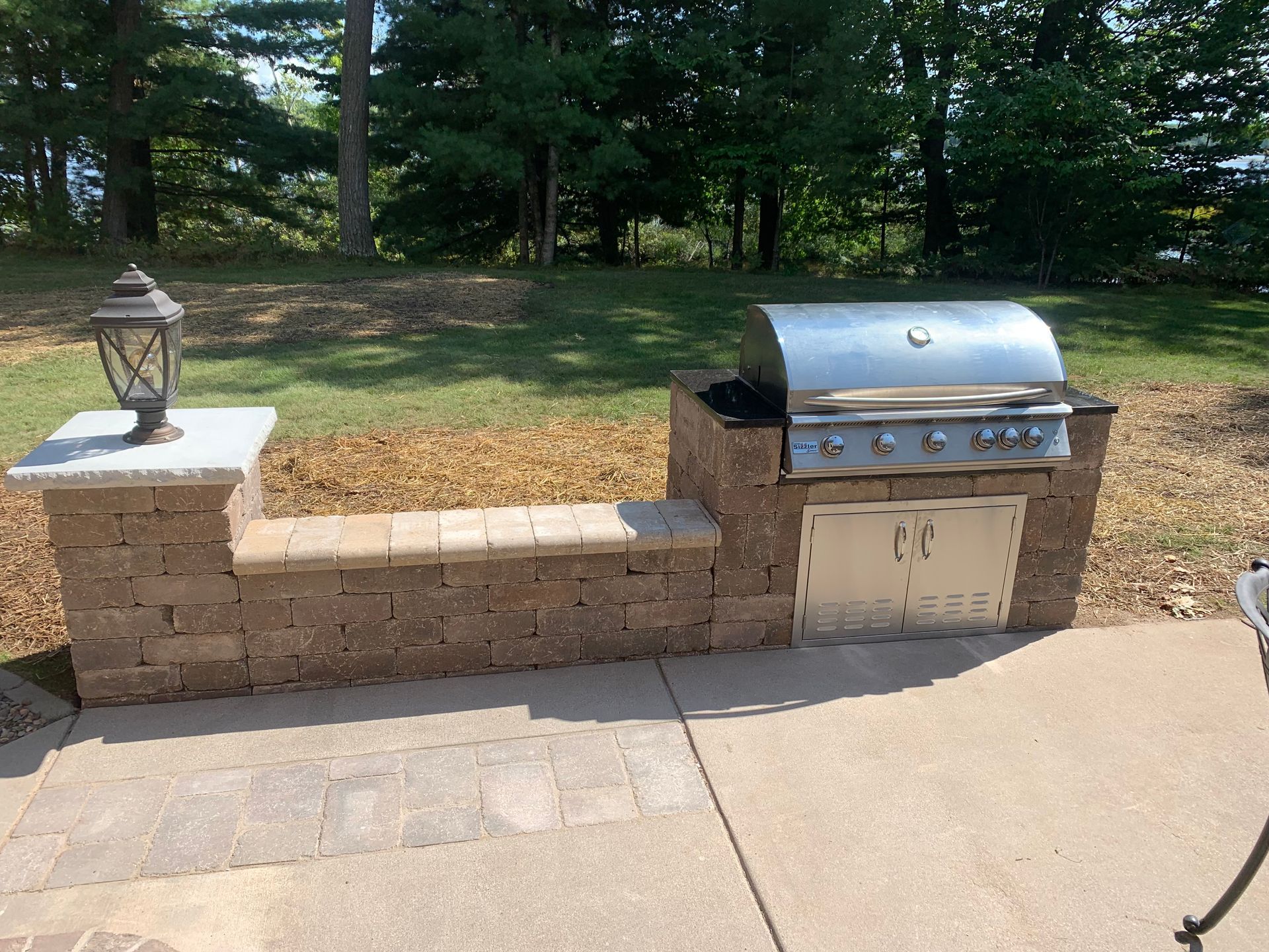 Stone outdoor grill and seating wall beside a paved patio in a wooded park setting