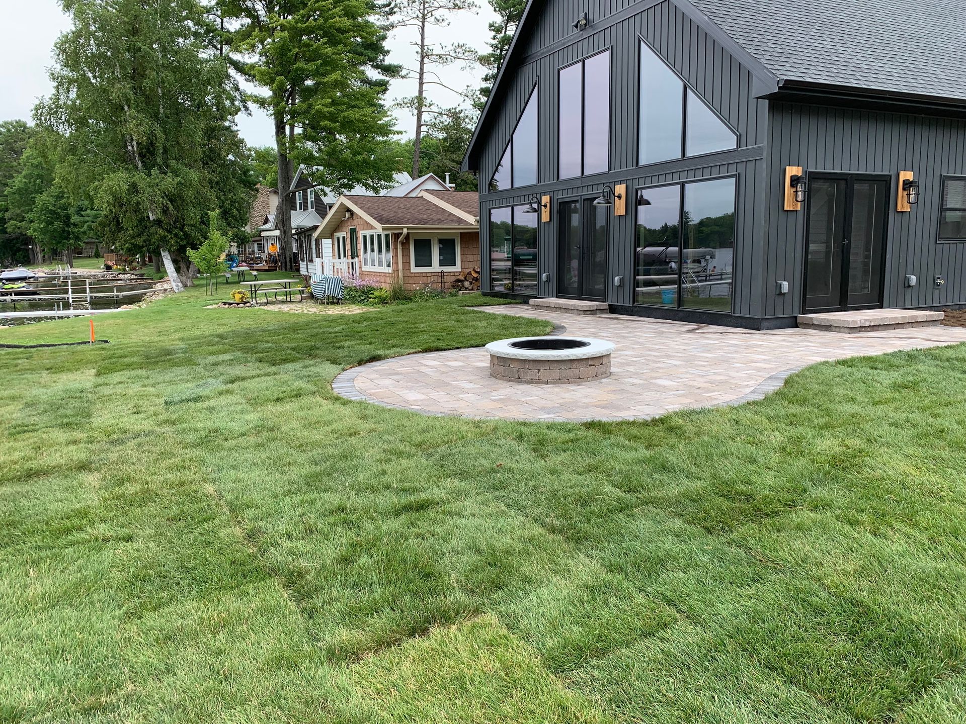 Modern black house with large windows, stone patio, fire pit, and a green lawn with trees nearby