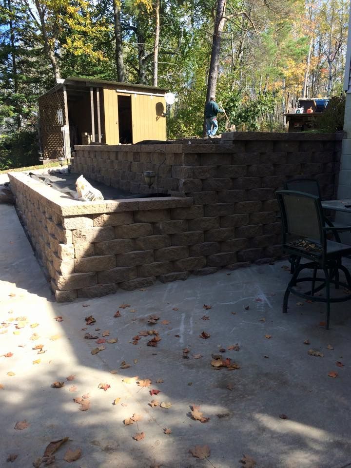 Stone patio with retaining wall, outdoor chair, and small shed in a wooded backyard