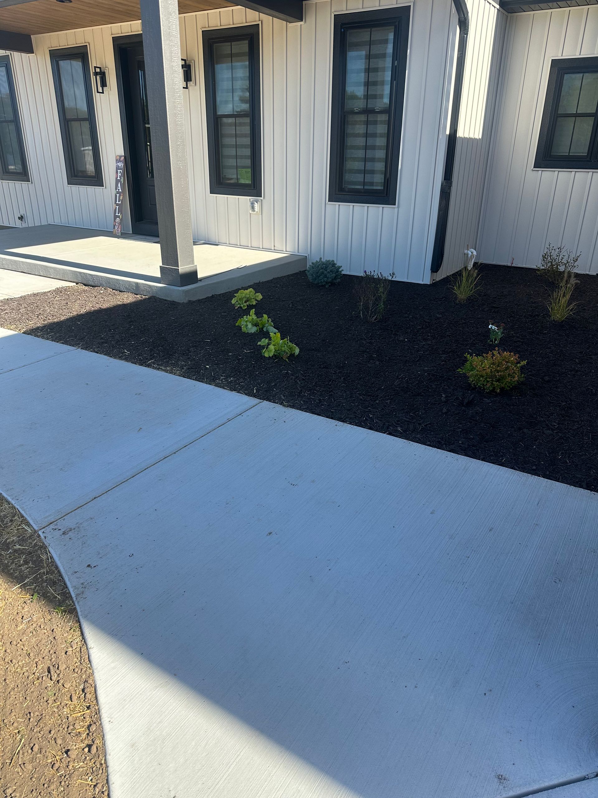 Concrete walkway beside a white house with black shutters and a small mulched planting bed