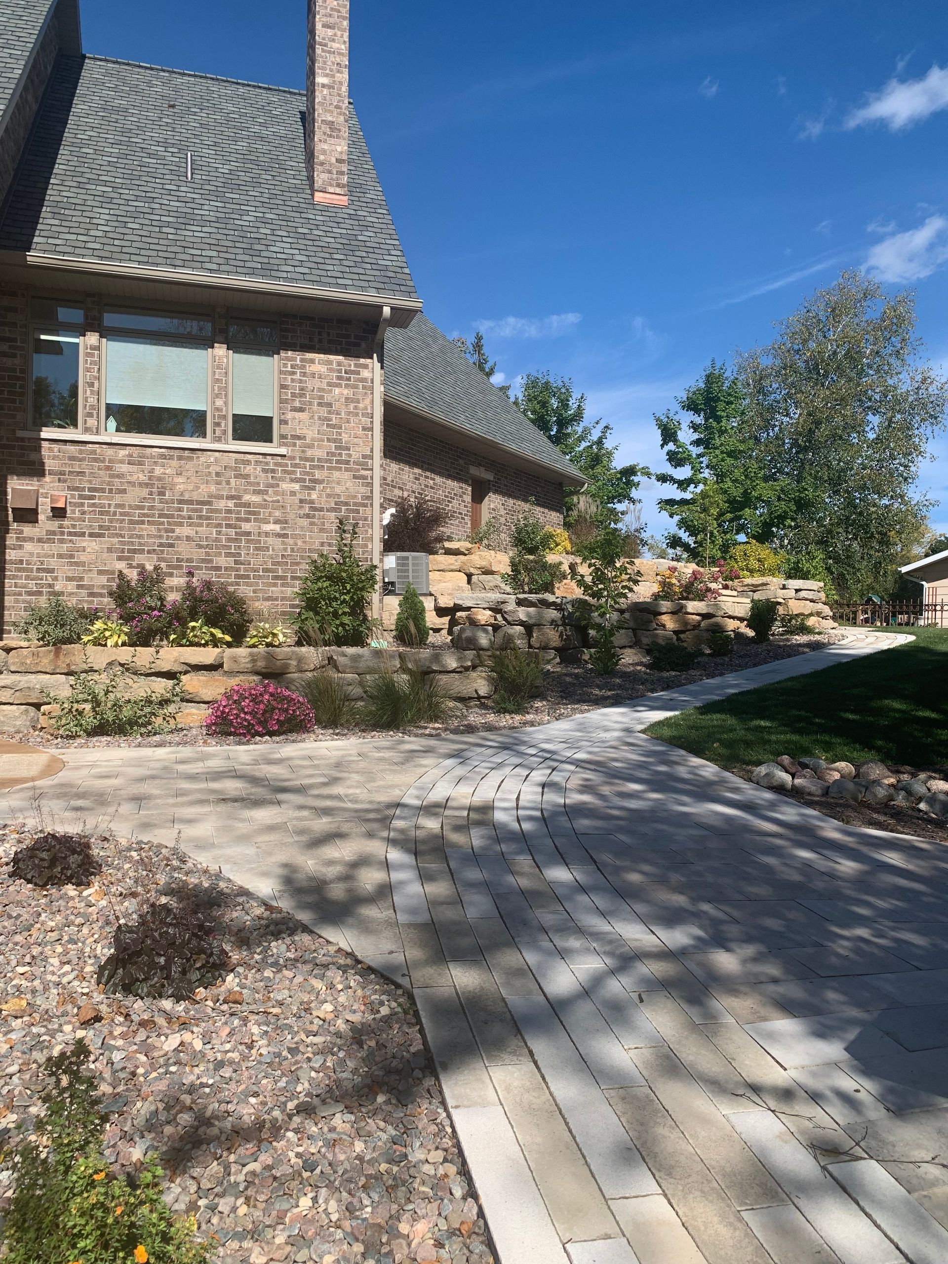 Stone house with blue-gray roof beside a curved paved walkway and landscaped garden under a clear blue sky