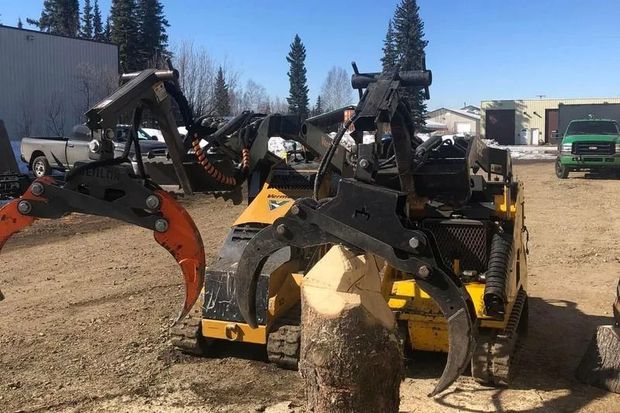 Two excavator attachments gripping a log in a dirt yard with trucks and equipment in the background