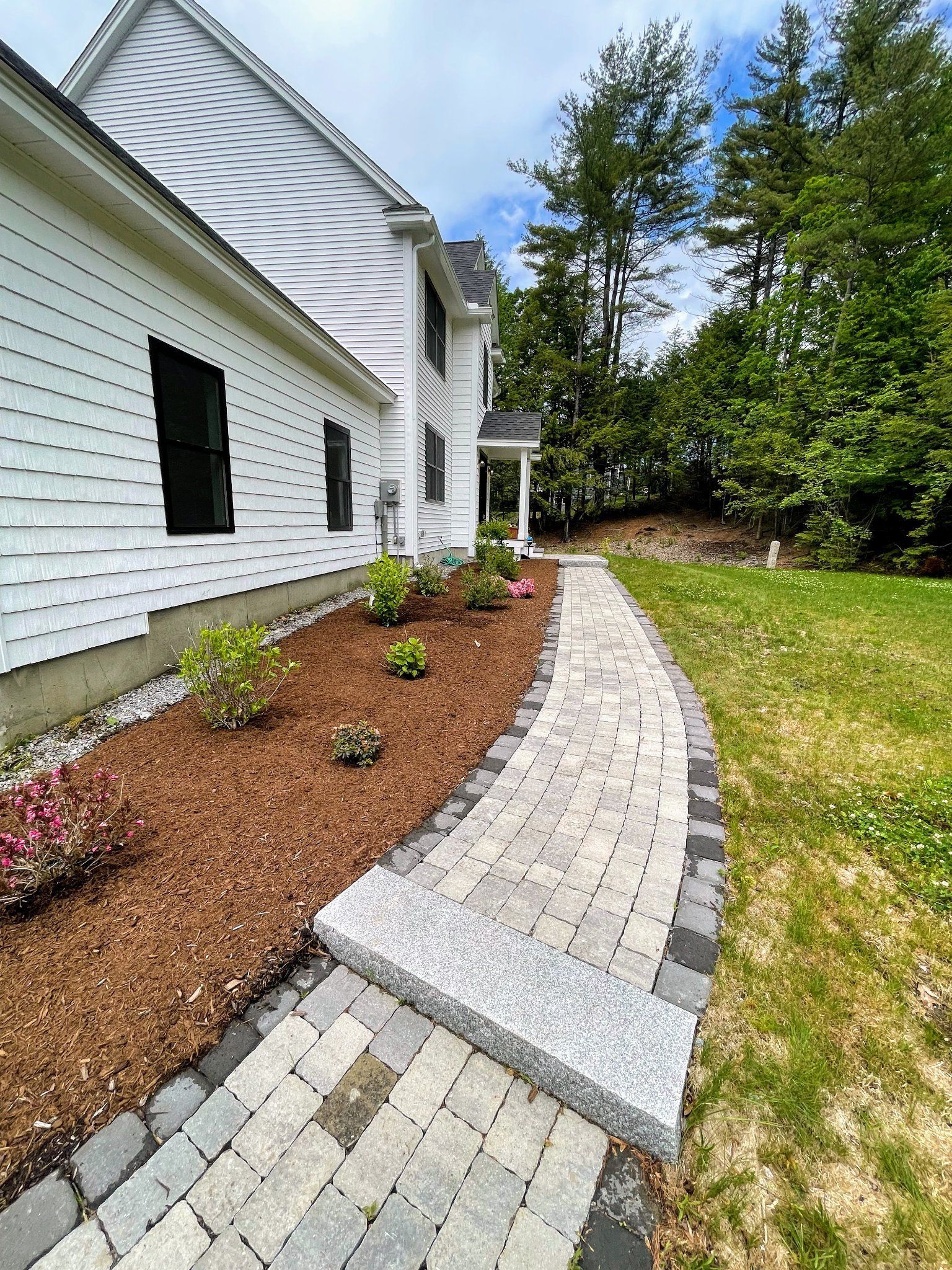 A brick walkway leading to a white house with trees in the background.