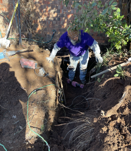 Person in a hole, digging with tools, near a brick wall and vegetation.
