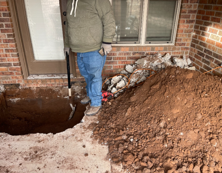 Person digging in a hole near a brick wall and a door, with dirt piled nearby.