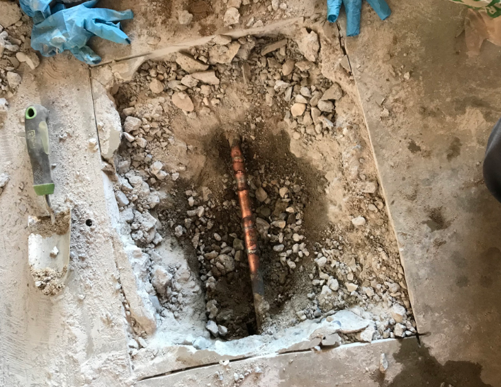 Hole in floor with exposed copper pipe, debris, and tools, likely plumbing repair.