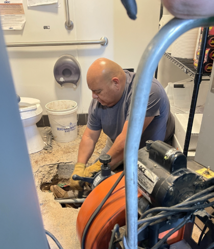A plumber repairs a pipe in a commercial restroom, using a drain snake.
