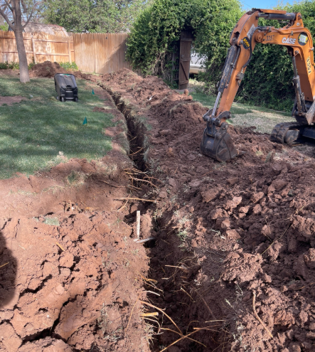 An excavator digs a trench in a yard, removing soil. Brown dirt and green grass are visible.