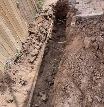 Trench dug alongside a wooden fence, revealing underground pipes. Dirt and roots are visible.