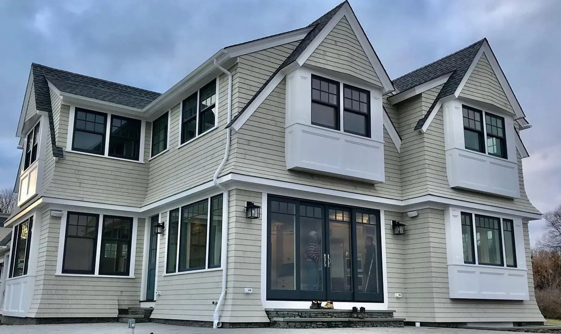 A two-story house with light siding and dark window frames under a cloudy sky. The house has multiple gabled dormers and a large glass door.