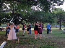 People dancing outdoors at a celebration under a large tree; a bride wears a pink dress.