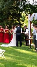 Wedding ceremony outdoors: Bride and groom exchanging vows, red-dressed bridesmaids, sunny, green lawn.