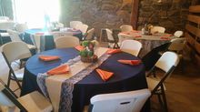 Round tables set for a gathering, navy tablecloths, orange napkins, white chairs in a rustic room.