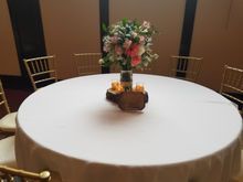 Round table with white tablecloth, floral centerpiece, gold chairs.