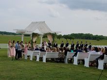 Outdoor wedding ceremony with bride and groom under a white arbor; guests seated on white benches, green field in background.