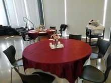 Tables set for an event; round, red tablecloths, black chairs.  Neutral room with blinds.
