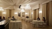 Elegant event space with round tables, chandeliers, and people seated. Neutral tones and draped ceiling.
