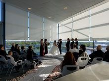 Wedding ceremony indoors. Bride and groom with wedding party, guests seated. Large windows with shades.