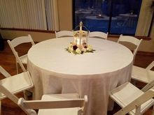 White-covered round table set for a gathering, with floral centerpiece and surrounding white chairs, near a window.