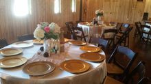 Round tables set for a gathering in a wooden-walled room. White linens, gold chargers, and floral centerpieces.
