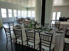 Elegant dining room with oval table set for a formal event; black chairs, white tablecloths, floral centerpieces.