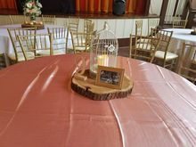 Round table with peach tablecloth, wooden centerpiece, birdcage, framed photo, gold chairs, and flowers.