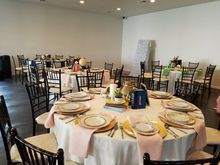 Tables set for an event with white tablecloths, gold-rimmed plates, pink napkins, and black chairs in a spacious room.