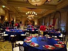Formal event room with round tables, blue tablecloths, red centerpieces, and chandeliers.
