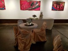 A wedding reception setup in an art gallery. Tables and chairs draped in cream fabric. Cake sits on a table. Art pieces on the wall.